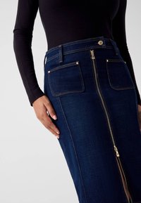 Dunkelblauer Denimrock mit einem Reißverschluss vorne, zwei aufgesetzten Taschen und goldfarbenen Beschlägen; kombiniert mit einem schwarzen Langarmshirt.