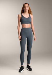 Szürke sportmellény és magas derekú leggings rugalmas anyagból. Simaság és varrás nélküli kialakítás jellemzi. Fekete sportcipő egészíti ki a megjelenést.