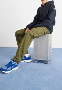 Blå sportskor med vita accenter, olivgröna cargobyxor och en mörk hoodie, sitter på en silverfärgad rullande box. Blå mattbakgrund.