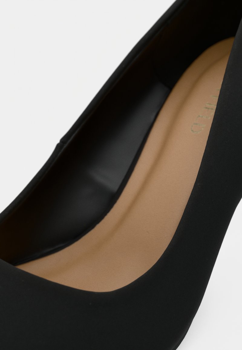 Gros plan d'une chaussure à talon noir montrant une semelle intérieure lisse couleur tan et un extérieur noir mat sur un fond uni.