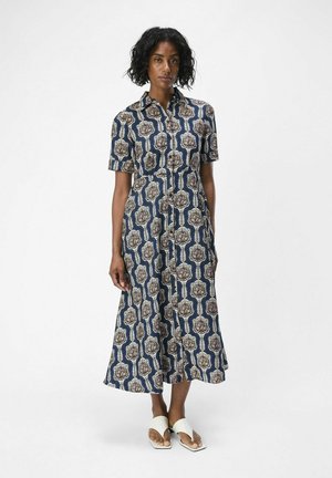 Vestido de manga corta con botones en azul marino y un patrón floral en beige y blanco. Silueta fluida, longitud hasta la mitad de la pantorrilla, combinado con sandalias blancas.