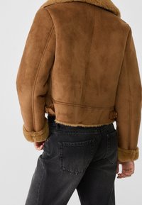Giacca in finta shearling marrone con design cropped, collettone oversize e polsini foderati in pelliccia. Abbinata a jeans grigi scuri con vestibilità rilassata.