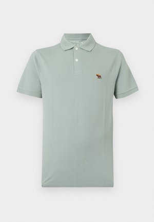 Ανοιχτοπράσινο πόλο με κοντά μανίκια και γιακά, δύο κουμπιά στο μπροστινό μέρος και μικρό κεντητό λογότυπο με μορφή άλκητα στο αριστερό μέρος του στήθους.