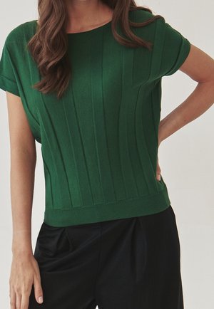Vrouw draagt een donkergroen geribd gebreid topje met korte mouwen, gecombineerd met zwarte, taillehoge broek, staat met één hand in de zij.