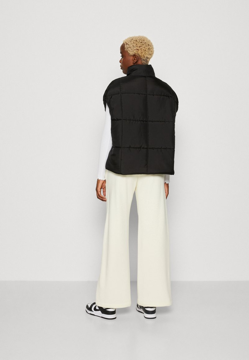 Black puffer vest with a high collar and quilted design, worn over a white long-sleeve top, paired with wide white trousers and black sneakers.