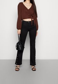 Brun kortärmad omslagstop med långa ärmar, svarta utsvängda jeans med knappelement, svart bucketbag och svarta sandaletter med remmar.