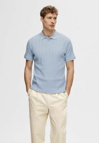 Svijetloplava kratkog rukava polo majica s vertikalnom rebrastom teksturom, s ovratnikom i opuštenim krojem, u kombinaciji s svijetlim hlačama.