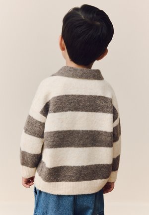 Małe dziecko z krótkimi ciemnymi włosami, ubrane w szary i biały sweter w paski oraz niebieskie dżinsy, stojące tyłem na jednolitym tle.