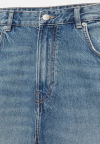 Jean en denim bleu en gros plan montrant la ceinture, le bouton avec le logo « Pull&Bear », les passants de ceinture, les poches avant et les détails de coutures contrastantes.