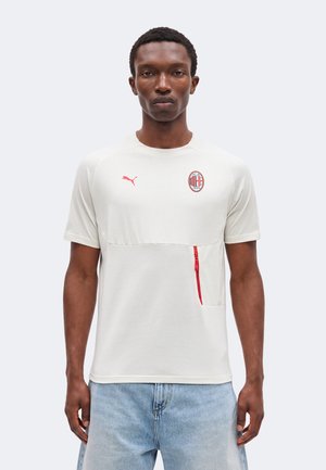 Uomo che indossa una maglietta bianca Puma AC Milan con dettaglio di cerniera rossa e jeans azzurri, in piedi davanti a uno sfondo neutro.