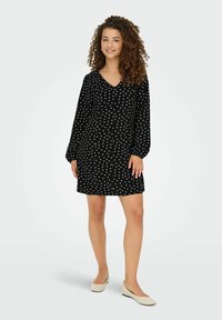 Jeune femme aux cheveux bouclés portant une robe noire à manches longues avec des motifs floraux blancs et des ballerines beiges, se tenant devant un fond uni.