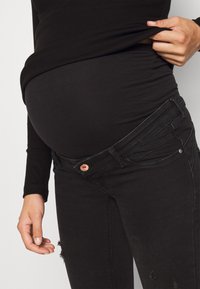 Svart graviditetstopp lager på lager över svarta slitna jeans, med en guldfärgad knapp och en mjuk, elastisk tyg för komfort.