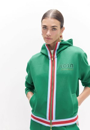 Mujer con sudadera verde con cremallera y rayas verticales rojas, blancas y azules y texto "LOLA CASADEMUNT", de pie con las manos en los bolsillos.