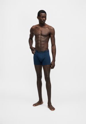Atletische donkerhuidige man staat op blote voeten, draagt marineblauwe boxershorts met een merkband, kijkt iets naar links.