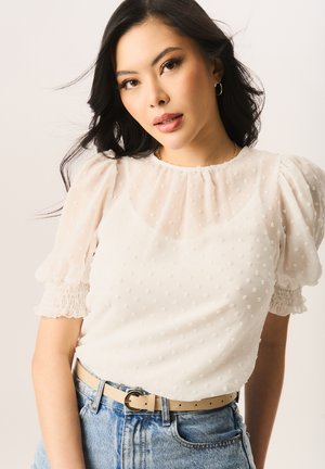 White blouse with puff sleeves and dotted texture, featuring gathered cuffs and a high neckline. Paired with high-waisted denim shorts and a light belt.