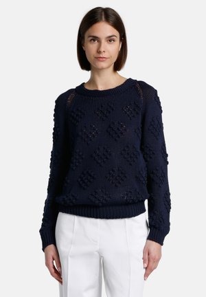 Navy blue knitted sweater with textured diamond patterns, round neckline, and ribbed cuffs and hem. Worn with white trousers.