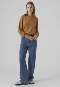 Pulover iz rjave volne z diamantnim vzorcem, v kombinaciji z modrimi jeans hlačami z visokim pasom in srebrnimi platform čevlji.