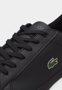 Μαύρο αθλητικό παπούτσι με μαύρα κορδόνια, με ανάγλυφο το όνομα της μάρκας στη γλώσσα και κεντητό πράσινο λογότυπο κροκόδειλου στο πλάι.