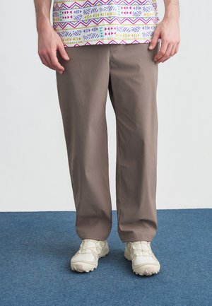 Mees, kes kannab helepruune pükse, valgeid tekstuuriga tossusid ja mitmevärvilise mustriga särki, seisab sinisel vaibal.