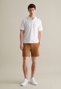 Polo blanco de manga corta con un bolsillo; combinado con pantalones cortos de ante color beige. Modelo vistiendo zapatillas blancas, de pie sobre un fondo neutro.