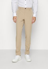 Beige színű, slim fit szabású, sima felületű kosztümnadrág, elöl gombos záródással és oldalsó zsebekkel, fehér sportcipővel párosítva.
