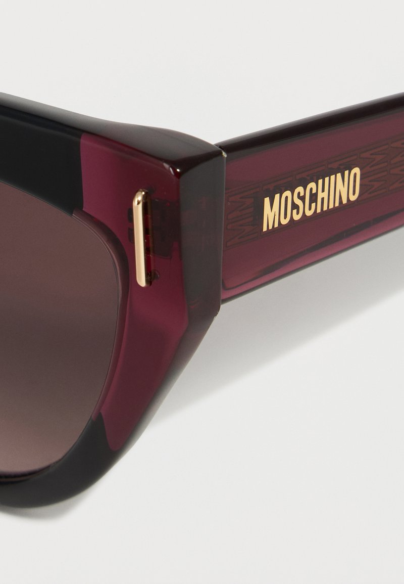 Close-up of dark red and black sunglasses with dark lenses and gold Moschino logo on the temple arm.