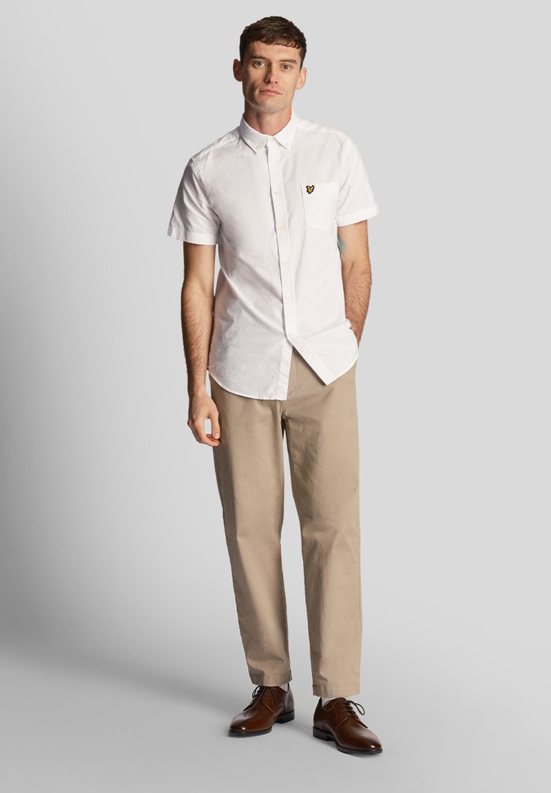 Camicia bianca a maniche corte con tasca sul petto e un piccolo logo, abbinata a pantaloni beige e scarpe in pelle marrone.