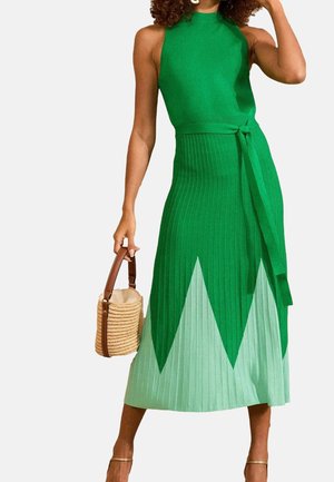 Femme en robe tricotée verte sans manches avec motif zigzag vert clair, tenant un sac à main en paille tressée avec des poignées en cuir marron.