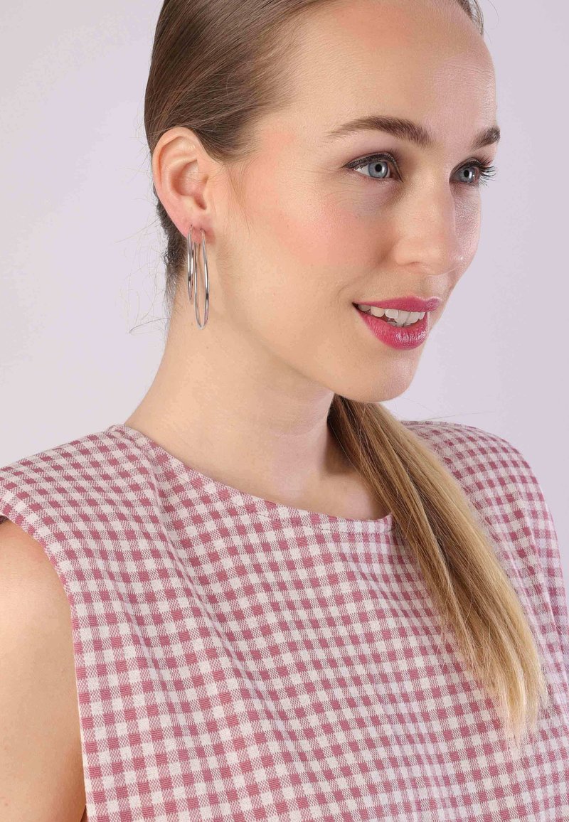 Lichtroze en crèmekleurig ruitjesjurk met een ronde halslijn en brede schouderbanden, aangevuld met grote zilveren hoepeloorbellen en steil haar.