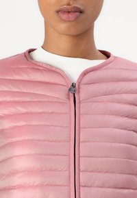 Casaco acolchoado rosa claro com design de quilt horizontal, gola alta e fecho frontal. Texto do logótipo visível ao longo da gola e textura suave.