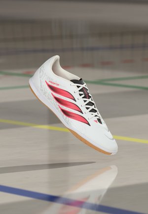 Chaussure de football en salle blanche avec des bandes rouges, des lacets noirs et une semelle en gomme, flottant au-dessus d'un sol de terrain de sport poli avec des lignes colorées.