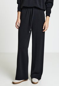 Schwarze, weit geschnittene Hose aus weichem Stoff mit elastischem Bund und Kordelzug. Kombiniert mit weißen Turnschuhen mit hellbrauner Sohle.