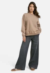 Frau mit langen, welligen Haaren, die einen lockeren beigen Pullover, weitgeschnittene dunkle Jeans und braune Sneakers trägt, steht mit einer Hand in der Tasche.