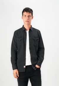 Zwarte denim shirt met knoopsluiting aan de voorkant, twee borstzakken en zichtbare stiksels. Draagt het over een wit T-shirt; model poseert tegen een lichte achtergrond.