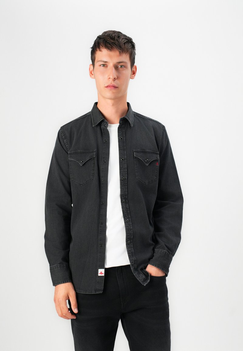 Czarna koszula dżinsowa z zapięciem na guziki z przodu, dwoma kieszeniami na piersiach i widocznymi szwami. Nosi ją na białej koszulce; model pozuje na jasnym tle.