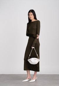 Olivzaļas krāsas garām piedurknēm piedurkņu kleita ar pieguļošu dizainu, kopā ar baltu rokassomiņu un smailu papēžu kurpēm, uz modeļa pret vienkrāsainu fonu.