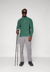 Green fleece zip-up top with black side panels, gray hiking trousers, and black hiking boots. Model holds trekking poles.