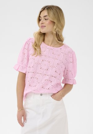 Blondynka w różowej koronkowej bluzce z krótkim rękawem i wzorami kwiatowymi oraz białe spodnie z wysokim stanem, z ręką w kieszeni, patrząca na bok.