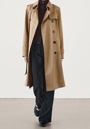 Person trägt einen beige doppelreihigen Trenchcoat über einem dunkelbraunen Hemd mit Knöpfen und lockeren schwarzen Hosen, läuft auf einem beigen Boden.