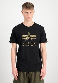 Juoda medvilninė marškinėlių su auksiniu ir kamufliažo Alpha Industries logotipo grafiką priekyje. Trumpomis rankovėmis ir apvalia iškirpte.