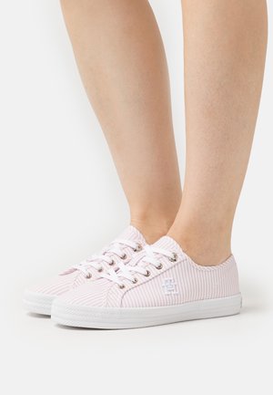 Persona que lleva zapatillas de lona de corte bajo en rosa claro y blanco con rayas, cordones blancos y suelas de goma blancas, de pie sobre una superficie blanca.