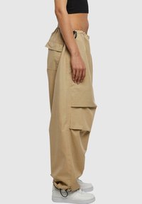 Pantalons cargo beiges en tissu léger, avec une taille élastique, des poches latérales et une coupe fuselée. Portés avec des baskets blanches.