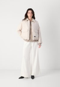 Jachetă bej deschis cu umplutură, dotată cu mari buzunare frontale și guler, combinați cu un pulover tricotat și pantaloni albi cu picioare largi.