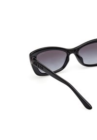Gafas de sol estilo cat-eye en negro con acabado brillante, lentes gris degradado y logotipo "GUESS" en relieve en el lateral. Diseño elegante con bordes redondeados.