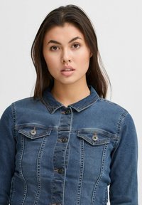Chaqueta de mezclilla en azul oscuro, con un diseño ajustado, cierre frontal con botones y dos bolsillos en el pecho. Presenta detalles de costura y un cuello.