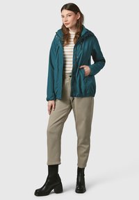 Giacca leggera color teal con cappuccio, chiusura con zip e polsini elasticizzati; abbinata a pantaloni beige e stivaletti neri con tacco.