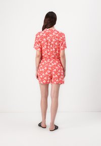 Rød floralmønstret romper med korte ermer, en tettsittende midje og shorts. Materialet ser lett ut, egnet for varmt vær.