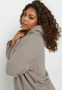 Yliresuinen beige huppari, jossa on teksturoitu kangas, suuri huppu ja ommeltu yksityiskohtia, malli, jolla on kiharat hiukset.