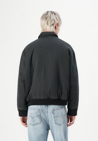 Persoon met kort blond haar, gekleed in een zwarte jas en lichtblauwe jeans, die met zijn rug naar de camera staat tegen een effen witte achtergrond.