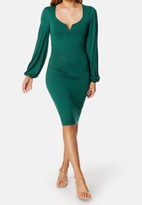 Robe verte à manches longues avec un décolleté en V, à la forme ajustée et en tissu texturé. Assortie à des sandales à talons transparents.